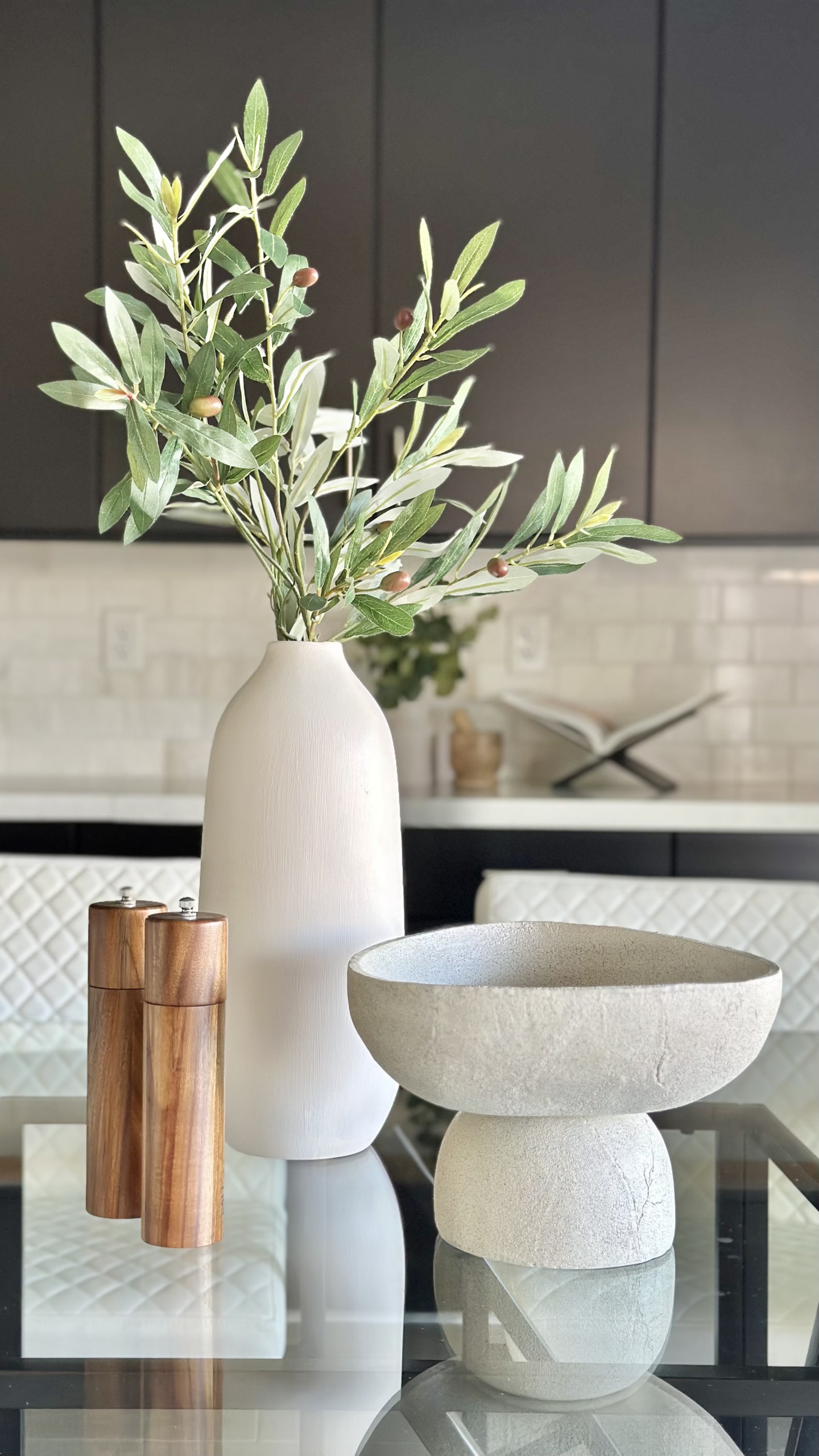 Staged kitchen counter vignette with olive branches in a white vase, pedestal bowl, and wood salt and pepper grinders — staging detail that elevates listing photos